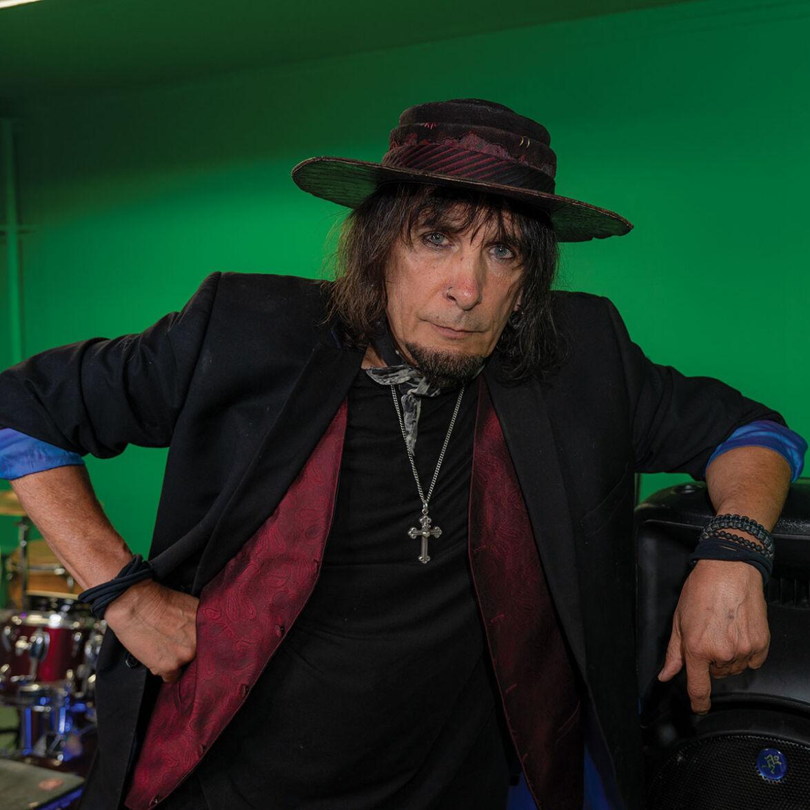 A man in. a brimmed hat and sports coat with the sleeves rolled rests his arm on a speaker in a green room.